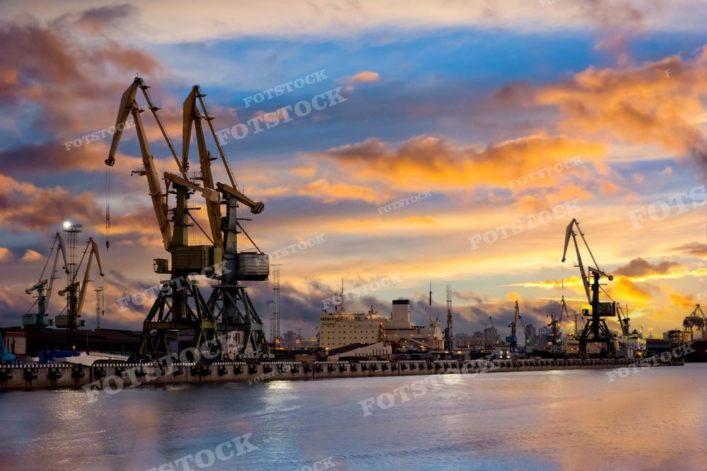 На фотографии изображен морской грузовой порт на закате. Видны морские грузовые терминалы и портовые сооружения для погрузки судов. Порт создает живописную картину на фоне заходящего солнца, напоминая о летнем вечере. На переднем плане видны погрузочные краны, готовые загрузить грузы на суда. 
Это изображение может быть использовано для иллюстрации темы морских портов, логистики и торговли, а также для привлечения внимания к красоте закатов и вечерних видов порта.